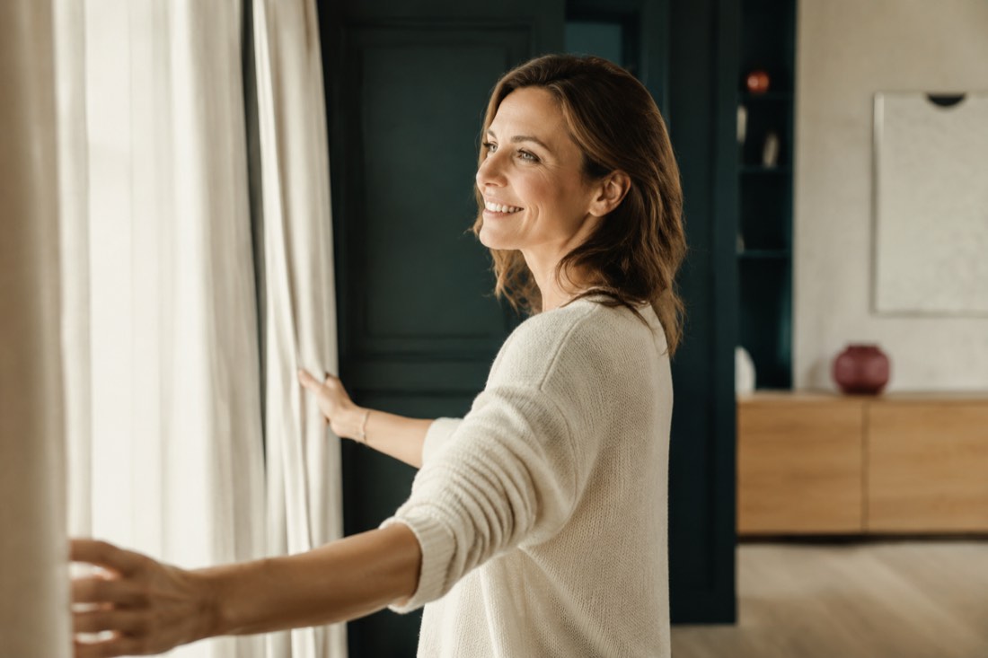 A smiling woman opening her curtains to the morning light